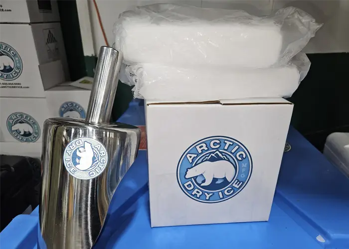 Bagged dry ice slices stacked on an Arctic Dry Ice shipping box beside a stainless steel scoop