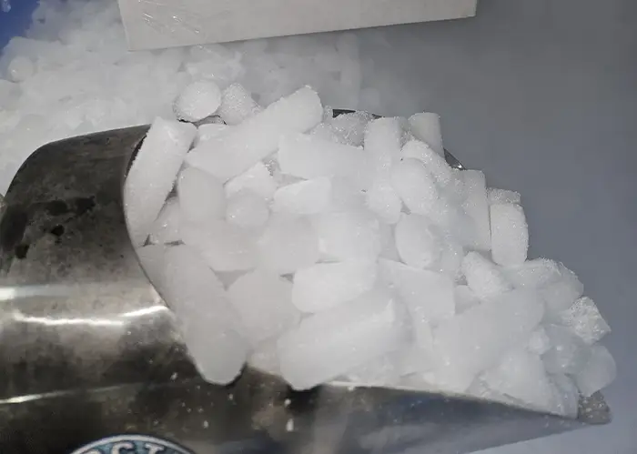 Scoop of 16mm dry ice pellets resting in a bin of loose pellets with visible vapor